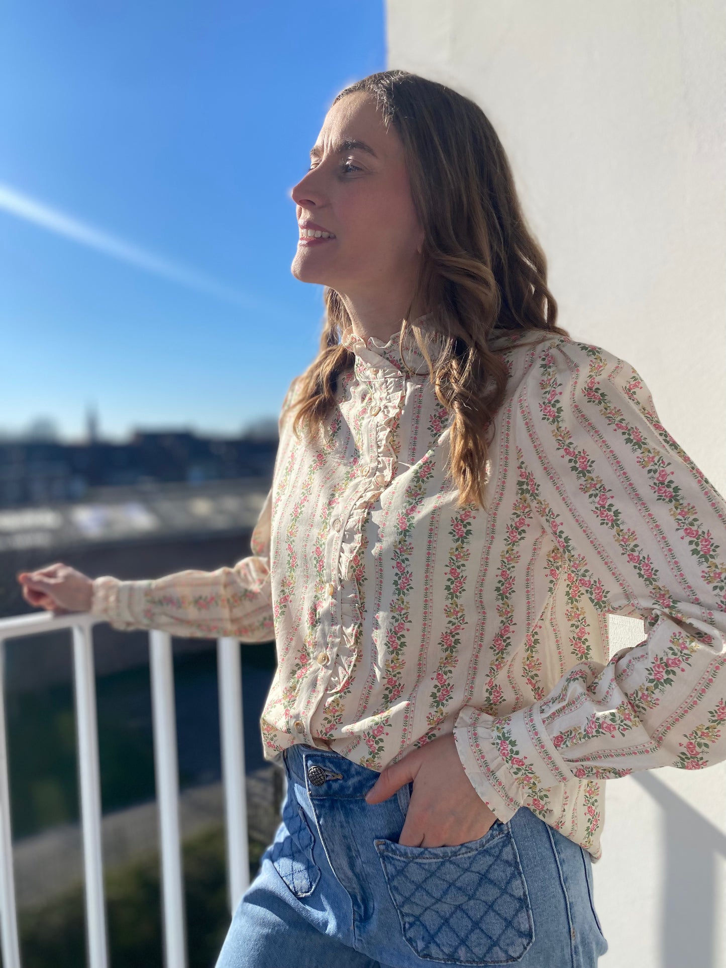 Femme portant la chemise Juliette beige à motifs fleurs roses, col à froufrous et jean bleu.
