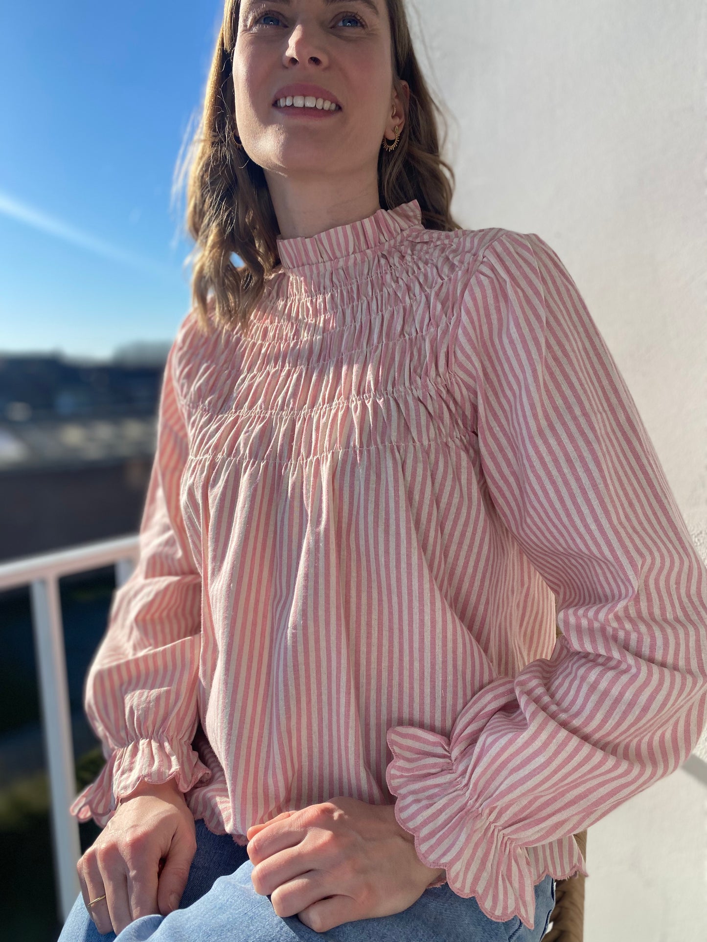 Femme assise portant la blouse Annette rose à rayures blanches avec col et buste smockés.