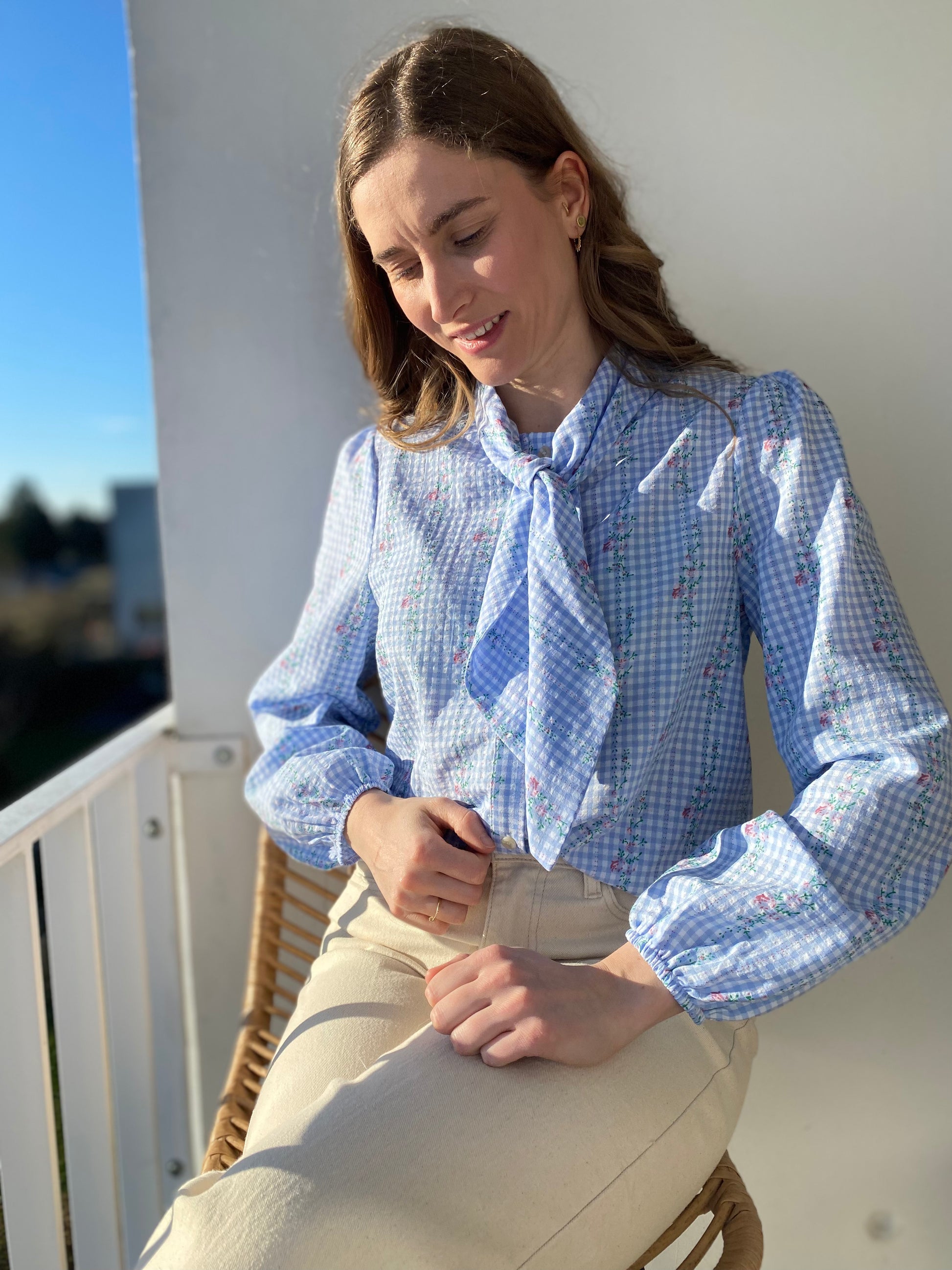 Femme portant une blouse bleue à carreaux vichy et fleurs avec un col noeud lavallière.